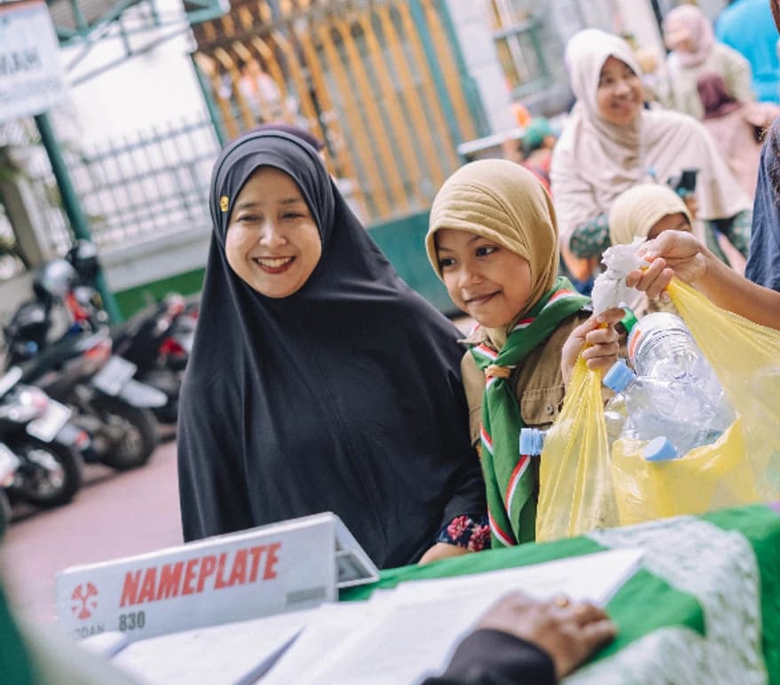Two collection members hand over bags of used plastic bottles at a street-side collection table