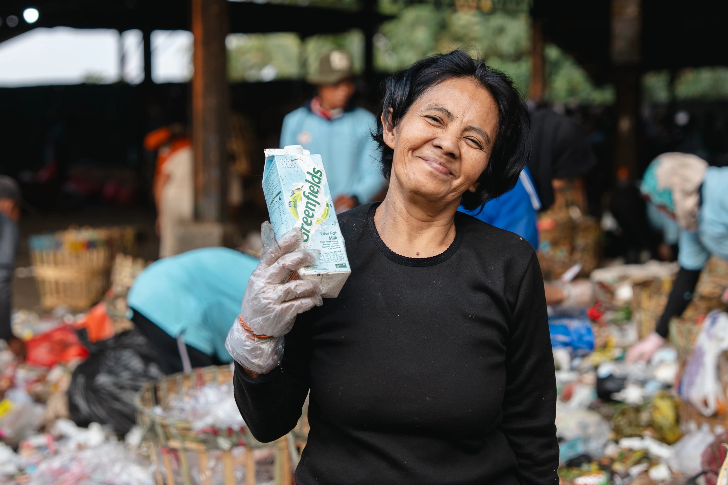 A smiling collector holding up an empty milk carton at a recycling site in Malang