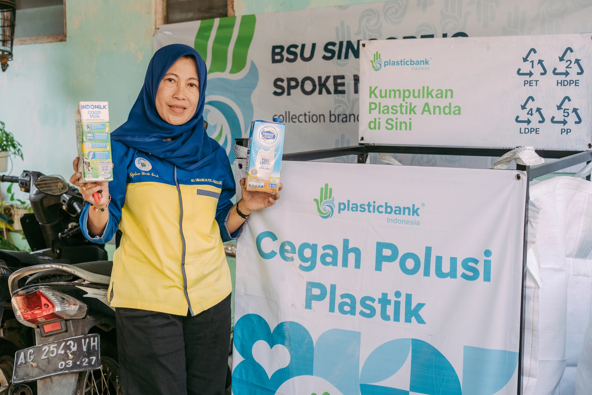 A waste bank member holding two milk cartons beside a Plastic Bank drop-off bin in Malang