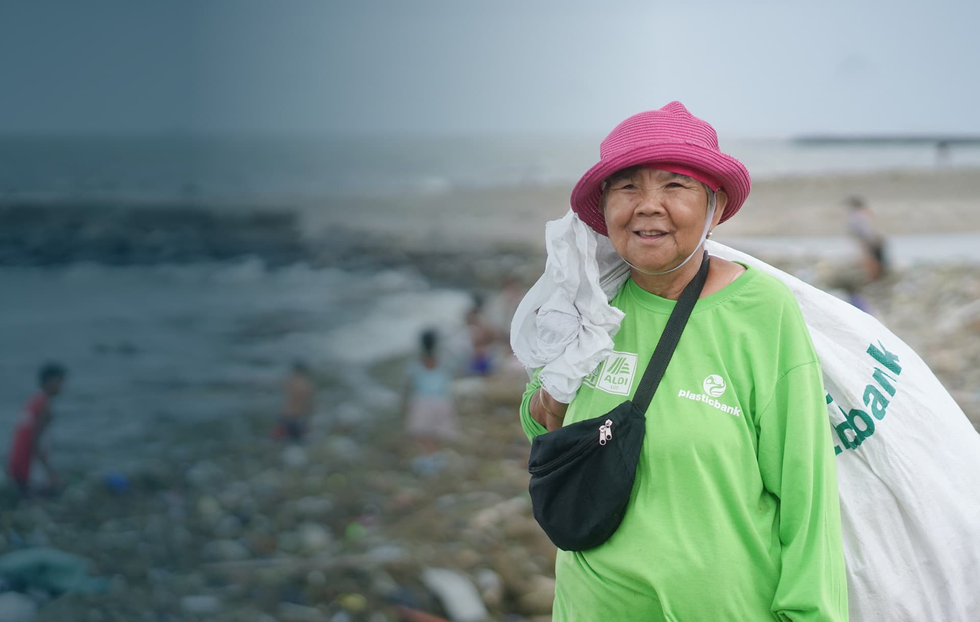 An elderly woman in a bright pink hat and a vibrant green Plastic Bank shirt smiles at the camera, holding a large white sack over her shoulder. She stands on a litter-strewn beach where other individuals can be seen in the background, engaged in a beach clean-up effort. Her presence highlights the importance of community involvement in environmental conservation.
