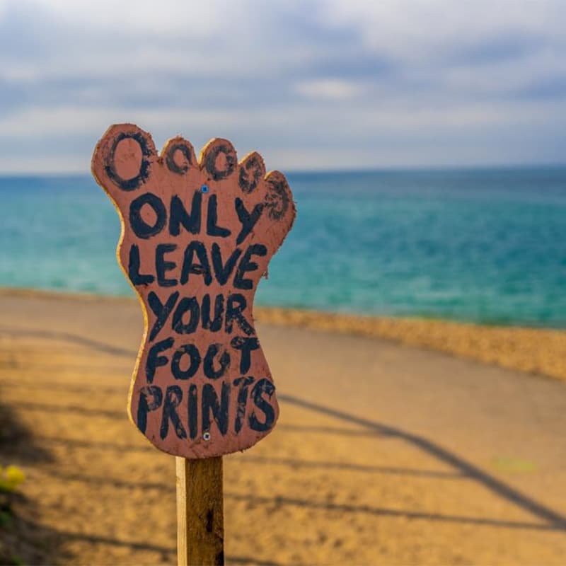 An eco-friendly sign shaped like a bare footprint stands on a sandy beach trail, with the phrase 'ONLY LEAVE YOUR FOOT PRINTS' written in bold letters, emphasizing the importance of reducing one's plastic footprint to preserve the natural coastal environment. The ocean in the background is in soft focus, highlighting the sign's message.