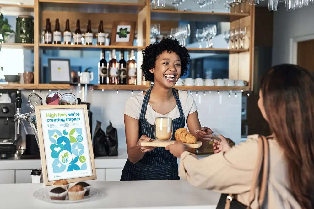 A barista with curly hair, wearing a black apron, smiles while serving a drink and croissants to a customer at a café.