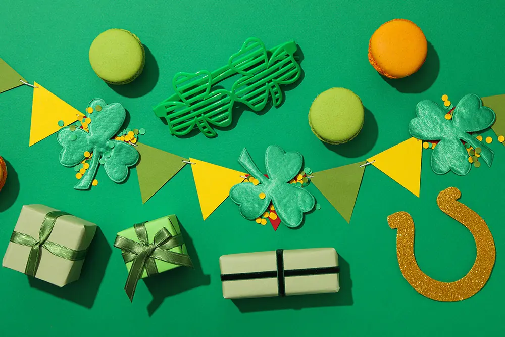 Overhead shot of St. Patrick's Day decorations including green shamrocks, green macarons, a gold horseshoe, green party glasses, gift boxes, and yellow and green pennant banners on a solid green background.