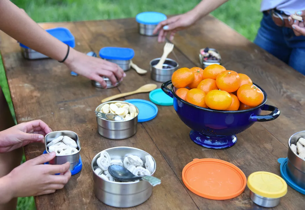 Hands reaching for stainless steel reusable food containers during an outdoor picnic with fresh fruits and snacks, promoting zero waste and plastic-free living